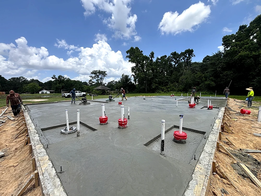 Crew finishing a freshly poured slab foundation, with plumbing stub-ups and beam lines visible